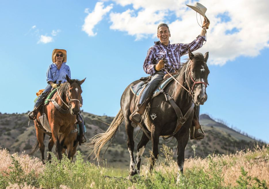 A man and a woman riding horses, the man holding his cowboy hat up in the air