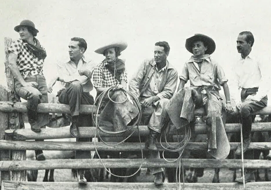 Old photo of a group of people sitting on a fence wearing riding clothes and holding lassoes